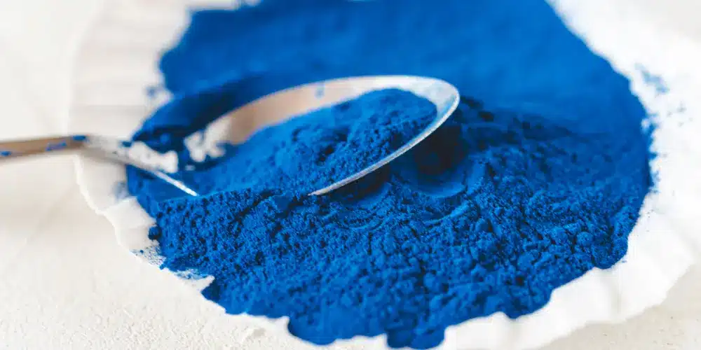 Une poudre d'un bleu éclatant est étalée sur une surface blanche, une cuillère en métal reposant dessus, créant ainsi un effet artistique contrasté et vibrant.