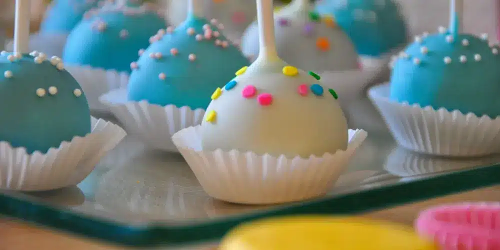 Gros plan sur des cake pops colorés, emballés dans du papier blanc, disposés sur un plateau. Certains sont bleu ciel avec des vermicelles blancs ; d’autres sont blancs avec des vermicelles colorés, créant une ambiance joyeuse et festive.