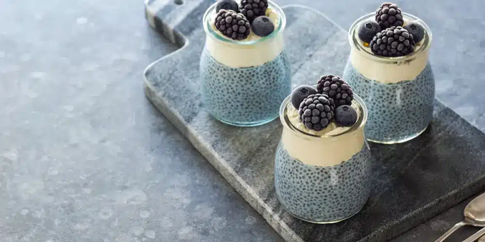 Trois pots en verre de pudding de chia garnis de crème, de mûres et de myrtilles, présentés sur une planche à découper en ardoise. La décoration est minimaliste et élégante.