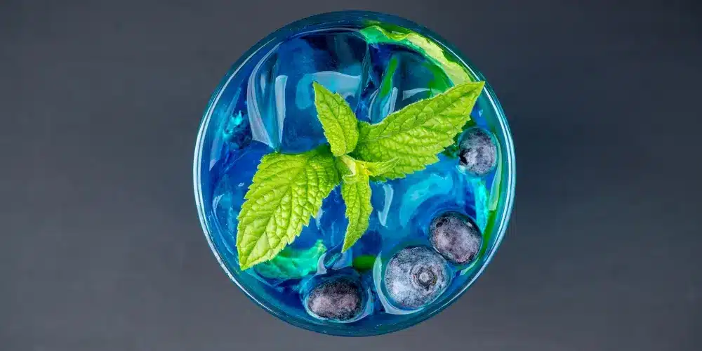 Une boisson d'un bleu éclatant, servie dans un verre, est ornée de feuilles de menthe fraîche et de myrtilles, sur un fond sombre, évoquant une ambiance estivale et rafraîchissante.