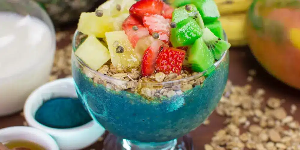 Un bol de smoothie coloré à base bleue, garni de granola, d'ananas frais, de fraises et de kiwi. À côté, des bols d'ingrédients sont disposés sur une surface en bois.
