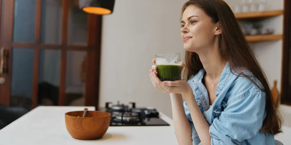 Une femme en chemise bleue est assise au comptoir d'une cuisine, souriante et tenant un verre de jus vert. Un bol en bois et une cuisinière sont visibles en arrière-plan.