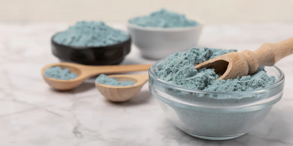 Trois bols et deux cuillères en bois remplis de poudre bleue sont posés sur une surface en marbre. La scène dégage une ambiance organisée, calme et naturelle.