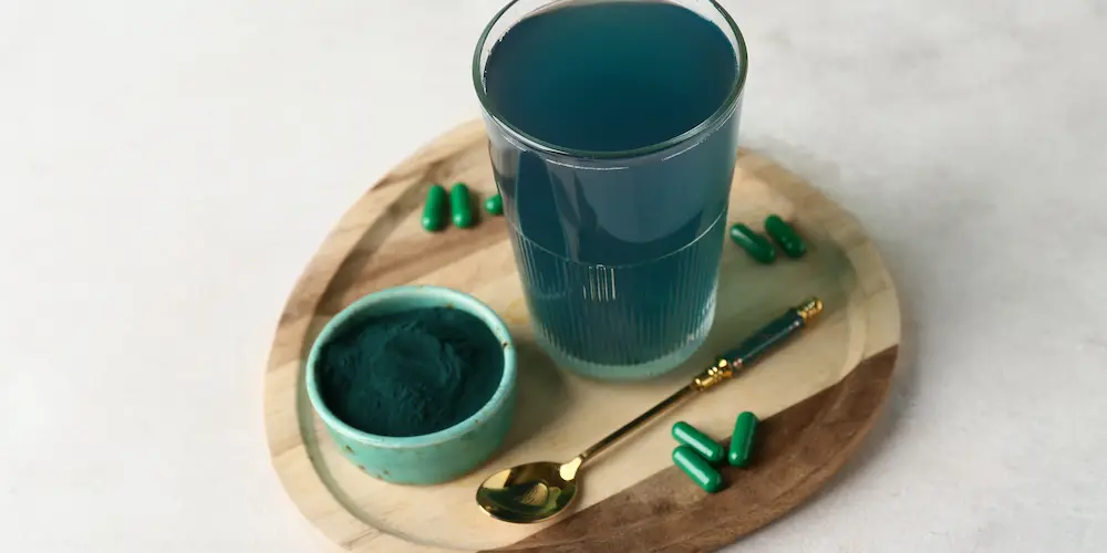 Un verre de liquide bleu, un petit bol de poudre bleue, une cuillère dorée et des capsules vertes sont disposés sur un plateau en bois sur un fond clair.