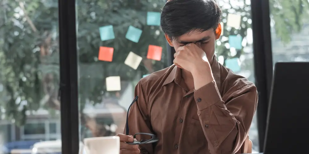 Un homme en chemise marron est assis à un bureau, des lunettes à la main, et se frotte les yeux, visiblement frustré. Des post-it colorés et un ordinateur portable sont visibles en arrière-plan.