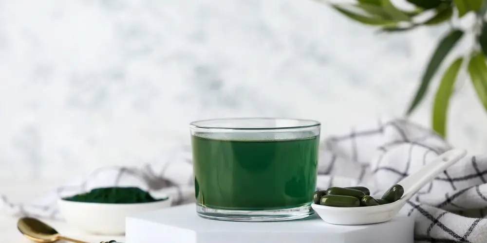 Un verre rempli d'un jus vert vif repose sur un bloc blanc, entouré de capsules vert foncé sur une cuillère et d'une plante feuillue, dégageant une impression de fraîcheur et de santé.