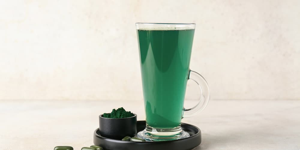 Un grand verre rempli d'une boisson verte à la spiruline repose sur un plateau noir. À côté, sur un fond clair, se trouvent un petit bol contenant de la poudre de spiruline et quelques capsules vertes.