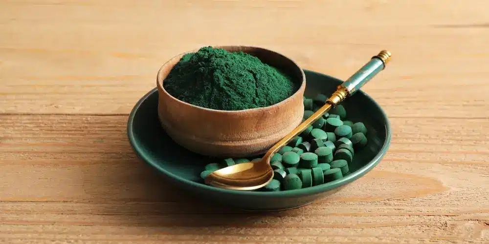 Un bol en bois rempli de poudre de spiruline verte repose sur une assiette verte, à côté de comprimés de spiruline verte. Une cuillère en or est posée sur l'assiette, le tout étant disposé sur une surface en bois.