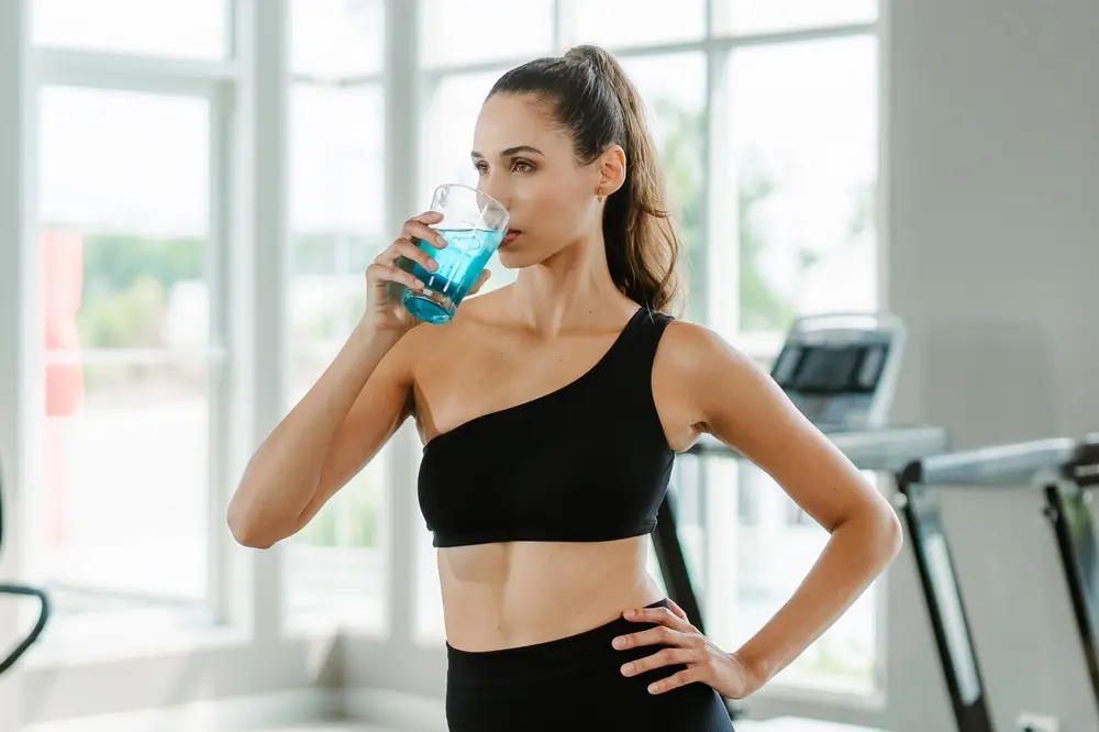Dans une salle de sport lumineuse, à côté de tapis de course, une femme vêtue d'un haut de sport noir asymétrique boit un liquide bleu dans un verre, affichant concentration et fraîcheur.