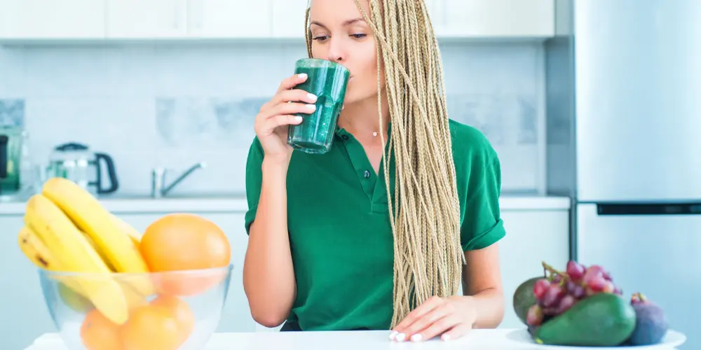 Dans une cuisine lumineuse, une femme aux longues tresses, vêtue d'une chemise verte, sirote un smoothie vert. Sur le plan de travail, un bol de bananes et d'oranges, ainsi qu'une assiette de raisins et d'avocats, évoquent un mode de vie sain et dynamique.
