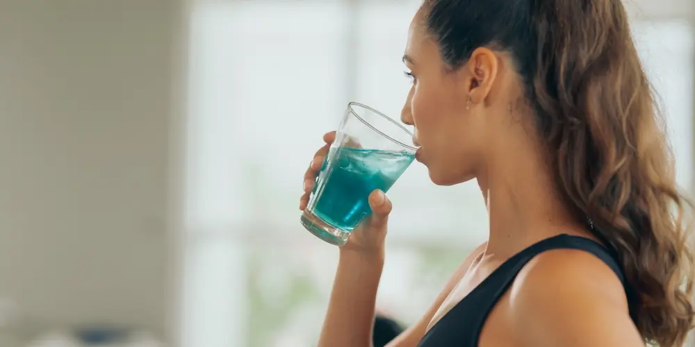Une femme aux cheveux attachés en queue de cheval, vêtue d'un débardeur noir, sirote une boisson d'un bleu éclatant dans un verre transparent. Elle paraît calme et concentrée dans un intérieur lumineux.