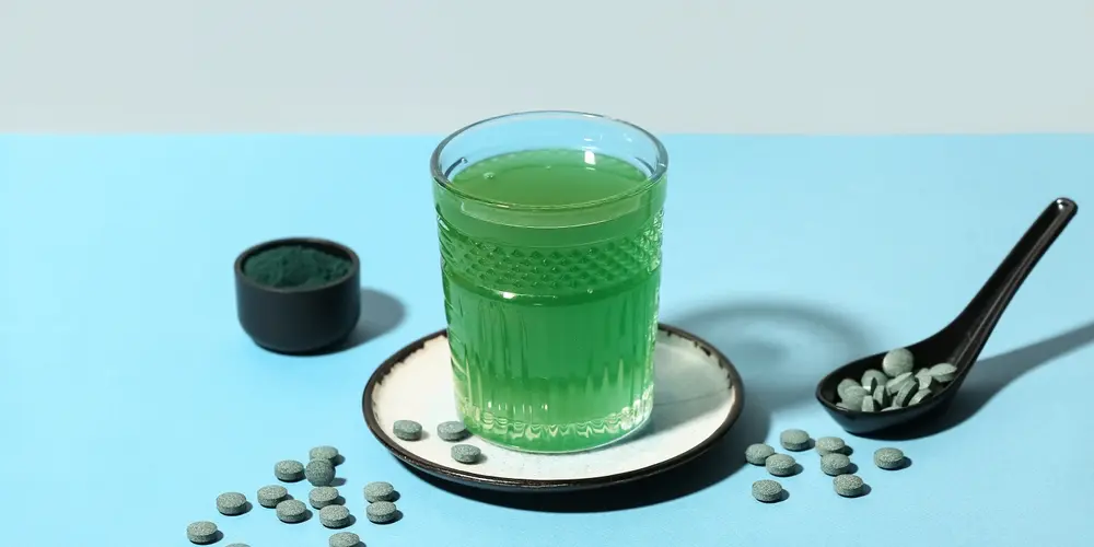 Un verre de jus vert vif sur une assiette blanche, entouré de comprimés de spiruline. Une cuillère noire avec des comprimés et un petit bol de spiruline en poudre à proximité.