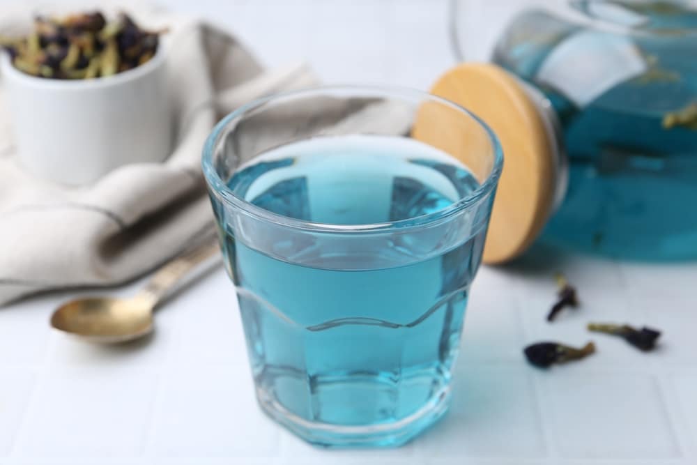 Un verre de thé aux fleurs de pois papillon d'un bleu éclatant repose sur une table. À proximité, des fleurs séchées, une cuillère et une théière en verre avec un couvercle en bois. Ambiance apaisante.