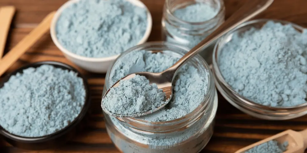 Des bols et des pots de différentes tailles, remplis de poudre bleu clair, sont disposés sur une surface en bois. Une cuillère prélève de la poudre dans un pot, créant une scène calme et ordonnée.