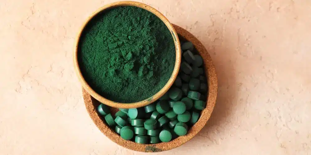 Un bol en bois contenant de la poudre verte repose sur un autre rempli de comprimés verts, le tout sur une surface texturée beige clair, évoquant un thème naturel et organique.