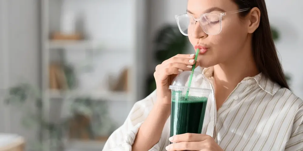 Dans un intérieur lumineux et flou, une femme à lunettes, vêtue d'une chemise à rayures, sirote un jus vert à la paille dans un gobelet transparent.