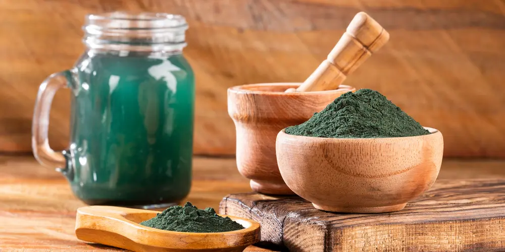 Des bols en bois remplis de poudre de spiruline verte sont posés sur une surface en bois. Un bocal en verre contenant une boisson à la spiruline verte est visible à l'arrière-plan. Ambiance naturelle et chaleureuse.