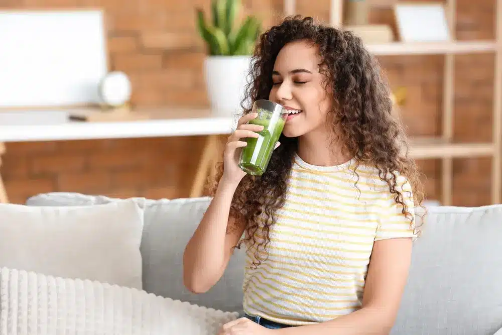 Une femme aux cheveux bouclés, vêtue d'une chemise à rayures, sourit en sirotant un smoothie vert sur un canapé confortable, avec une plante et des étagères en arrière-plan.