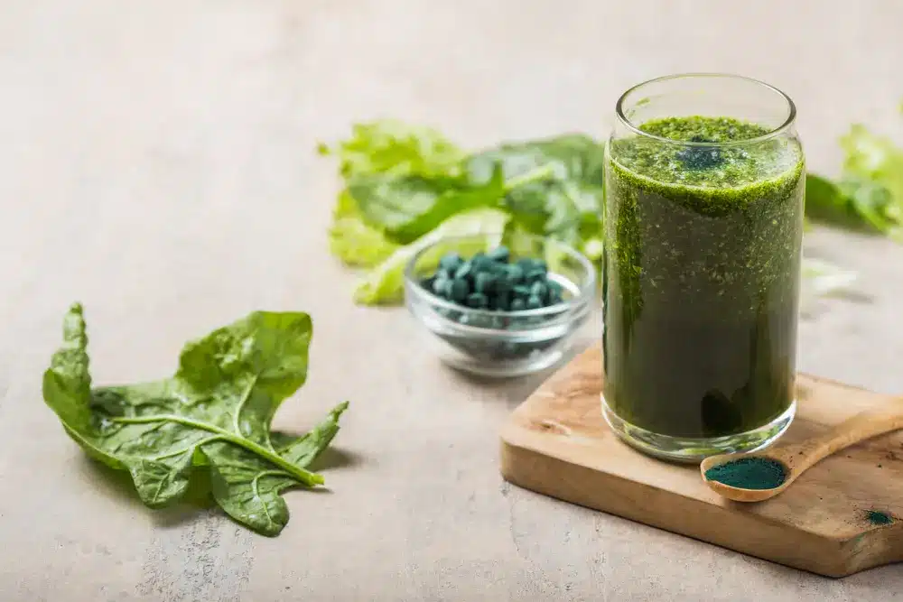 Un grand verre de smoothie vert posé sur une planche en bois, entouré de feuilles d'épinards frais et de comprimés de spiruline. Une cuillère en bois contenant de la poudre de spiruline est disposée à proximité.