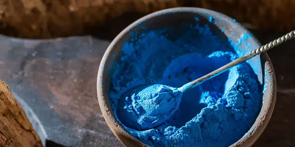 Un bol en bois rempli d'un pigment bleu vif, avec une cuillère posée à l'intérieur. Le fond est une surface sombre et texturée, créant une ambiance riche et artistique.