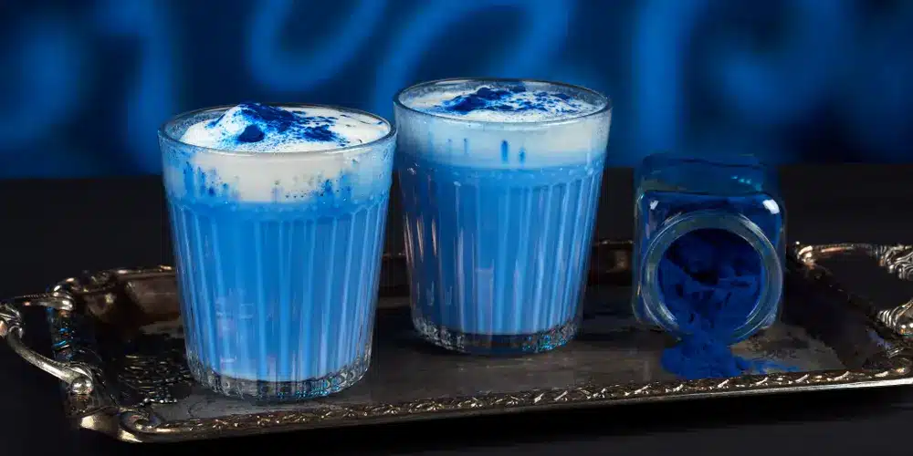 Deux verres de latte d'un bleu éclatant, coiffés d'une mousse onctueuse, posés sur un plateau argenté, à côté d'un pot renversé de poudre bleue, sur un fond bleu flou.