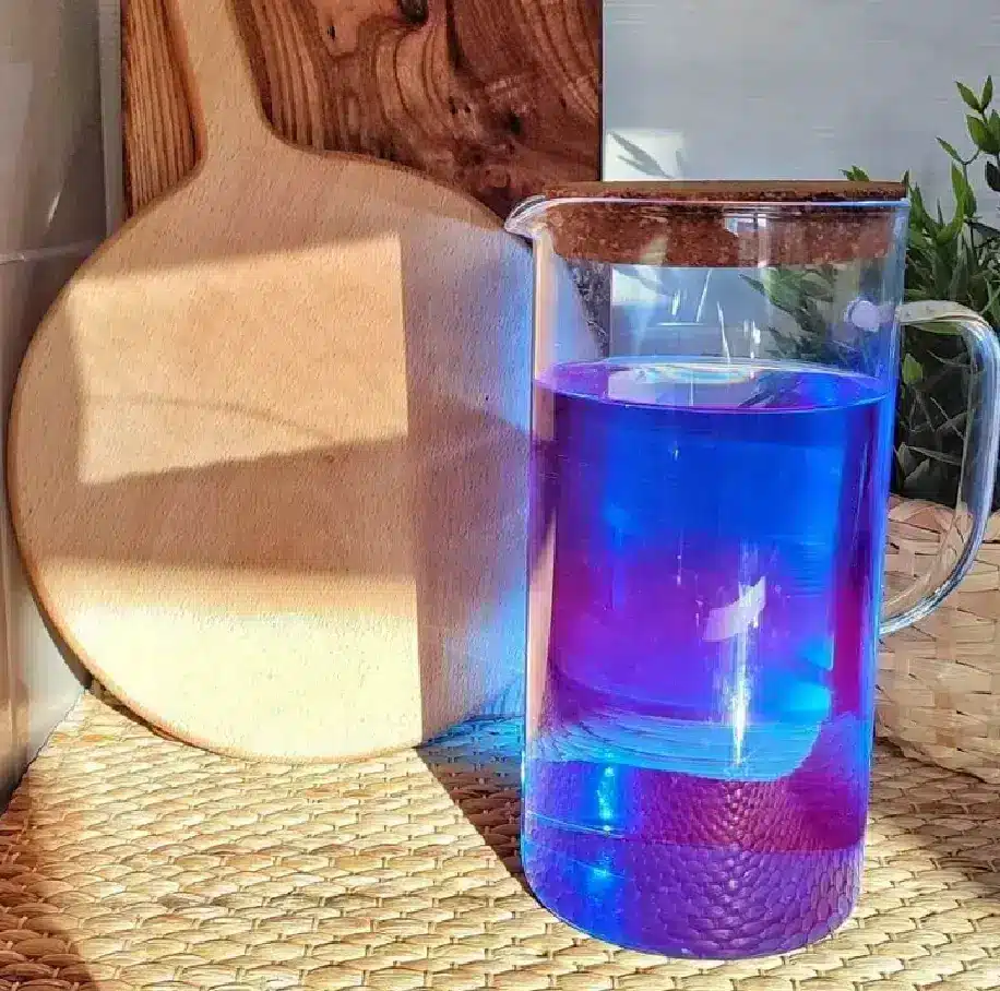 A clear glass pitcher with vibrant blue and purple liquid sits on a woven mat. Sunlight casts a shadow on a wooden cutting board nearby, creating a warm atmosphere.