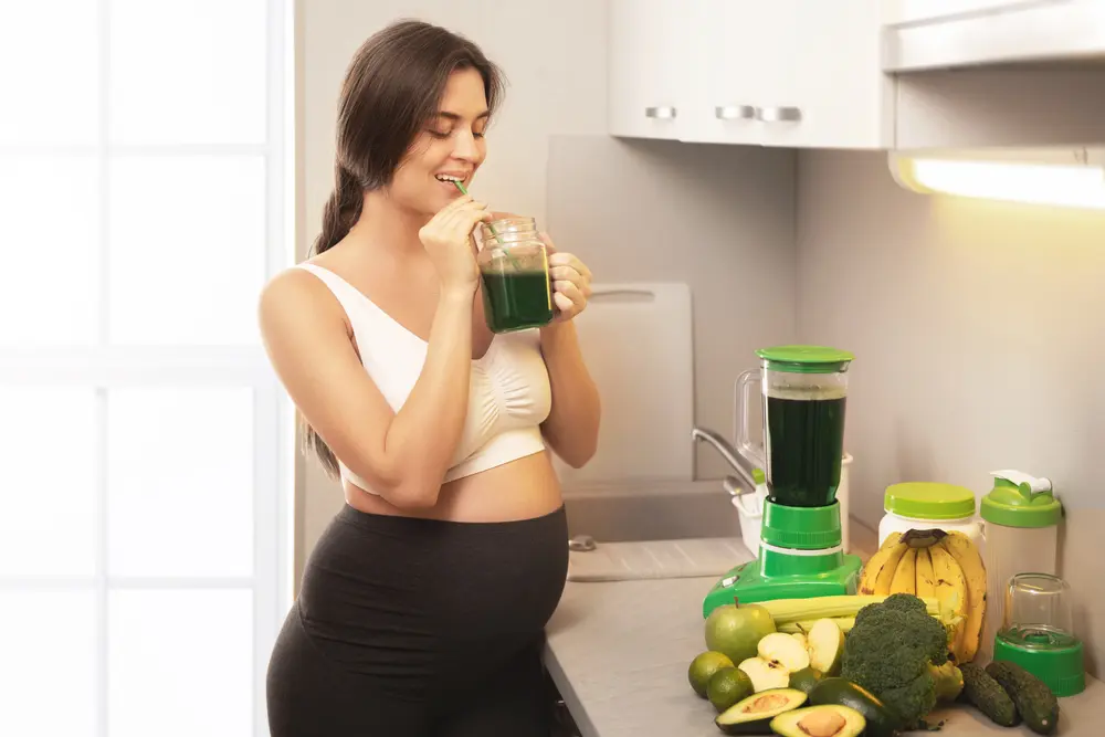 Jeune et heureuse femme enceinte buvant un smoothie vert sain à la maison.
