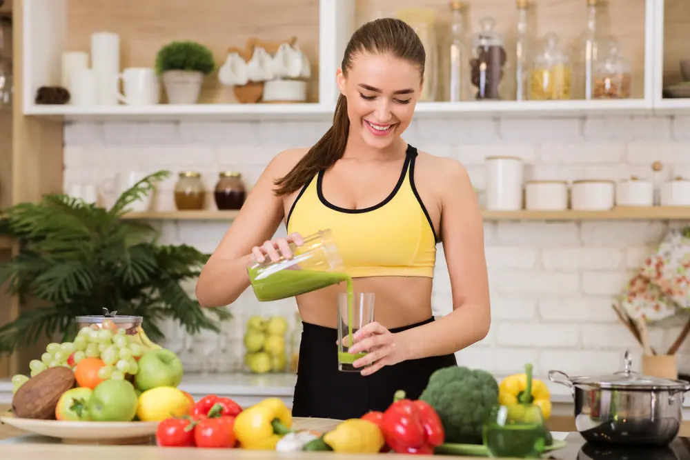 Une jeune femme en forme prépare un jus détox vert à la maison après son entraînement, vêtue de sa tenue de sport.