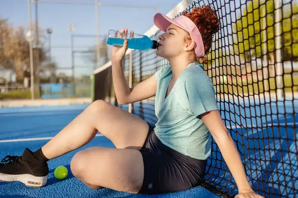 La jeune joueuse de tennis est assise sur le court, appuyée contre le filet, buvant une boisson énergisante bleue pour se rafraîchir pendant une pause dans son entraînement.