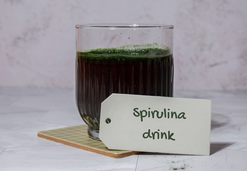 Un verre de boisson à la spiruline d'un vert foncé est posé sur un sous-verre. Une étiquette portant la mention « Spirulina drink » est placée devant, évoquant une ambiance saine et naturelle.