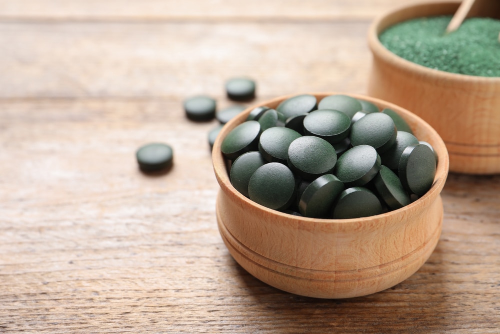 Bols de comprimés et de poudre de spiruline sur une table en bois, espace pour du texte.