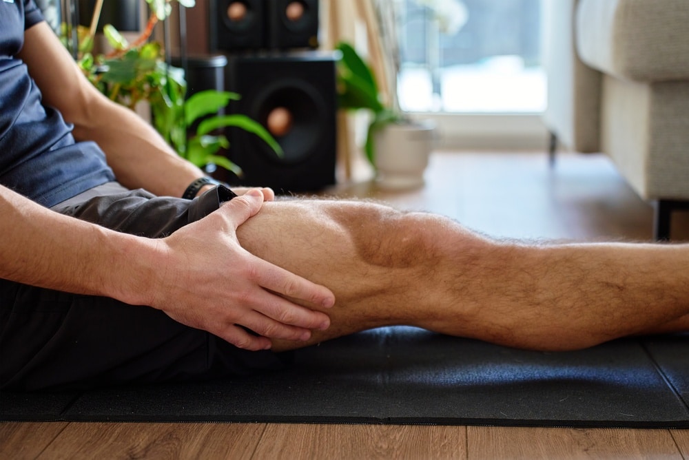 Homme se tenant le muscle de la jambe tout en étant assis sur un tapis de fitness, à l'intérieur. Concept de récupération musculaire, de soin du corps, de fatigue liée à l'entraînement et de rééducation.