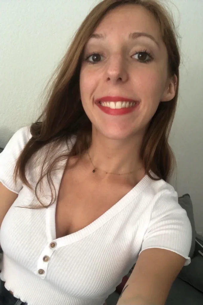 Jeune femme souriante aux cheveux châtains et au rouge à lèvres rouge porte un haut blanc à boutons et un collier fin.